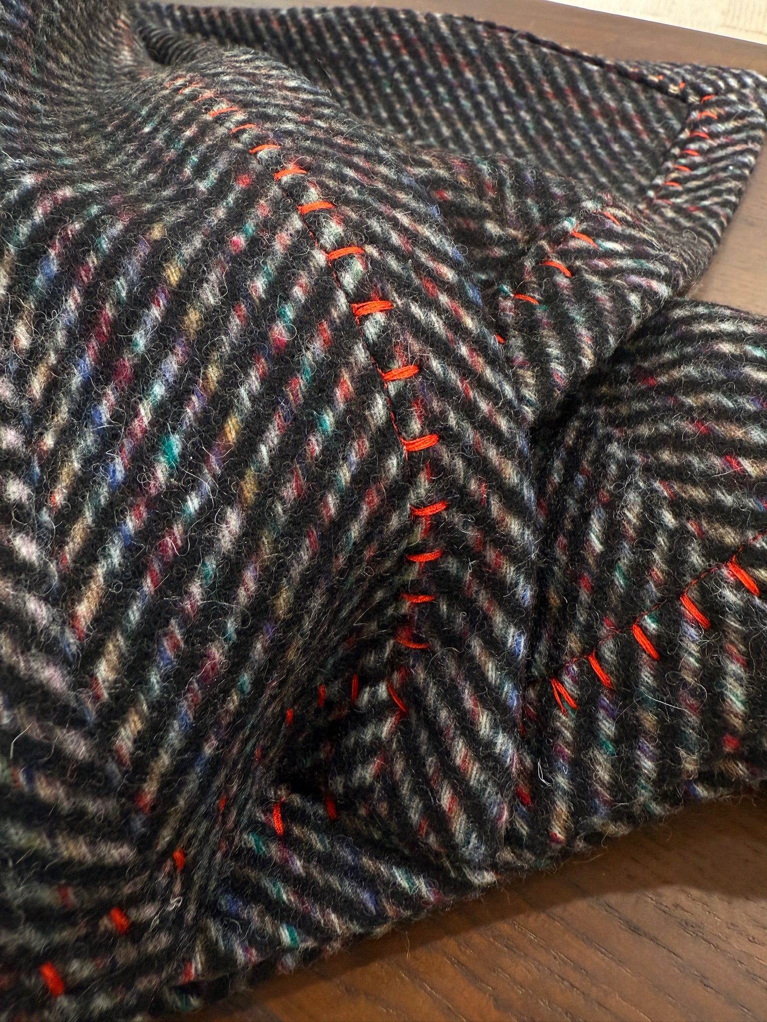 A close-up of a charcoal herringbone wool throw showcasing its soft felted texture, multicolor flecks, and bold red whipstitch edging. The detailed shot highlights the craftsmanship and cozy depth of the fabric.