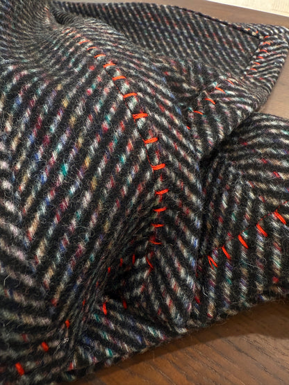 A close-up of a charcoal herringbone wool throw showcasing its soft felted texture, multicolor flecks, and bold red whipstitch edging. The detailed shot highlights the craftsmanship and cozy depth of the fabric.