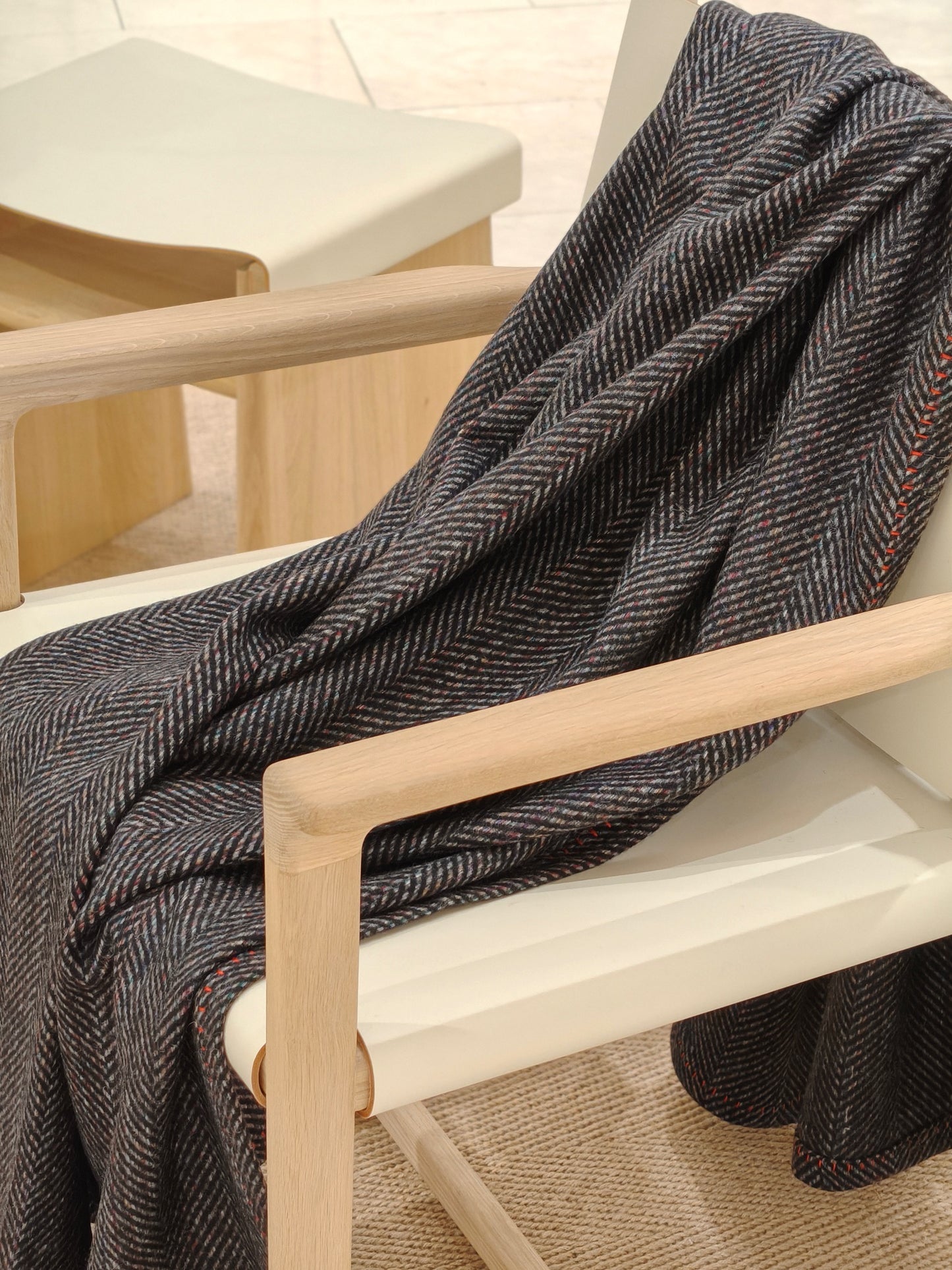 A charcoal herringbone wool throw draped over a modern wooden chair, showing its soft drape, multicolor speckled texture, and red whipstitch border. A warm, inviting lifestyle shot ideal for home décor presentation.