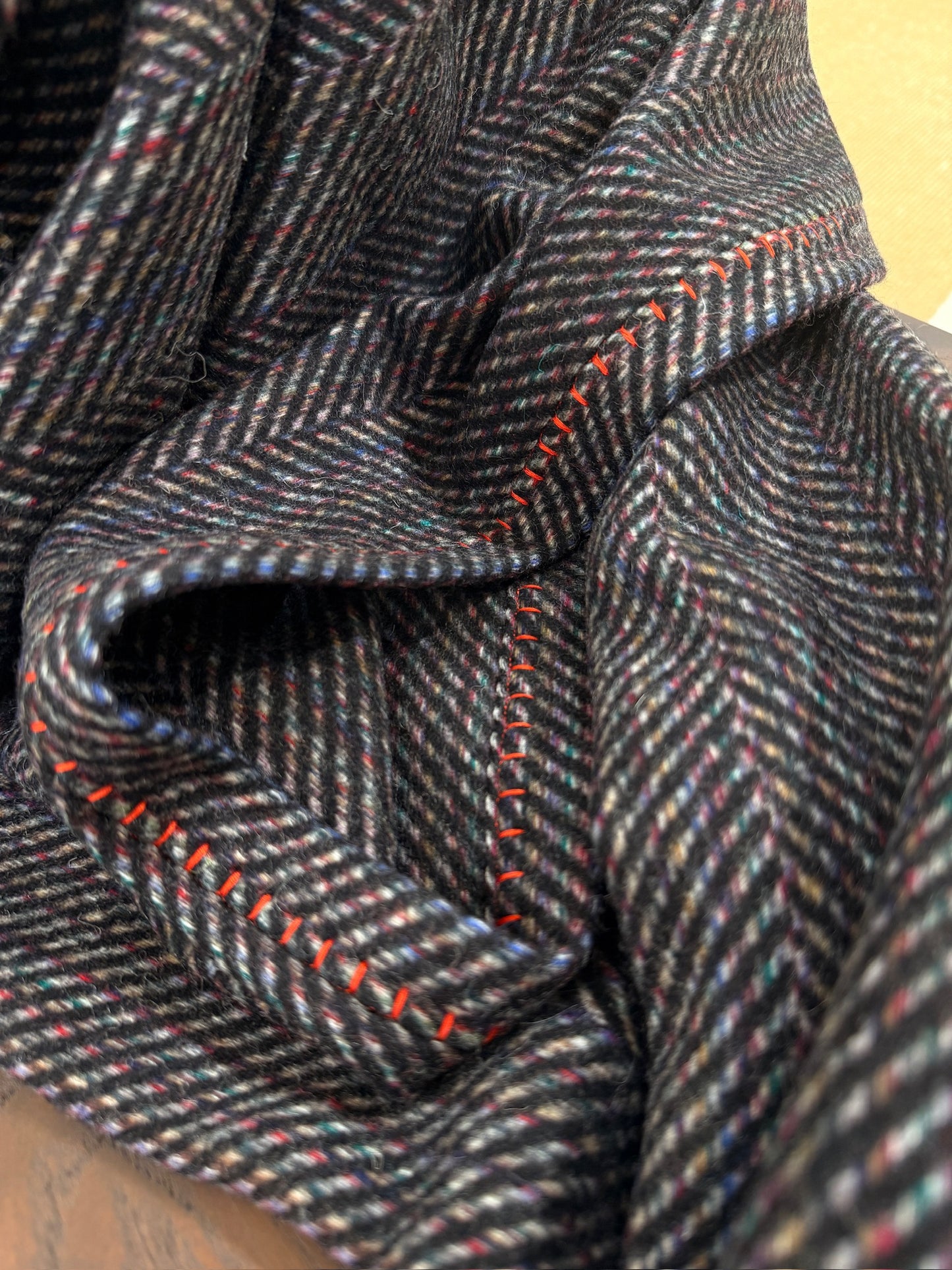 A detailed view of the wool throw’s herringbone weave with colorful yarn accents and vivid red hand stitching. The curved folds emphasize thickness, warmth, and artisanal finishing.