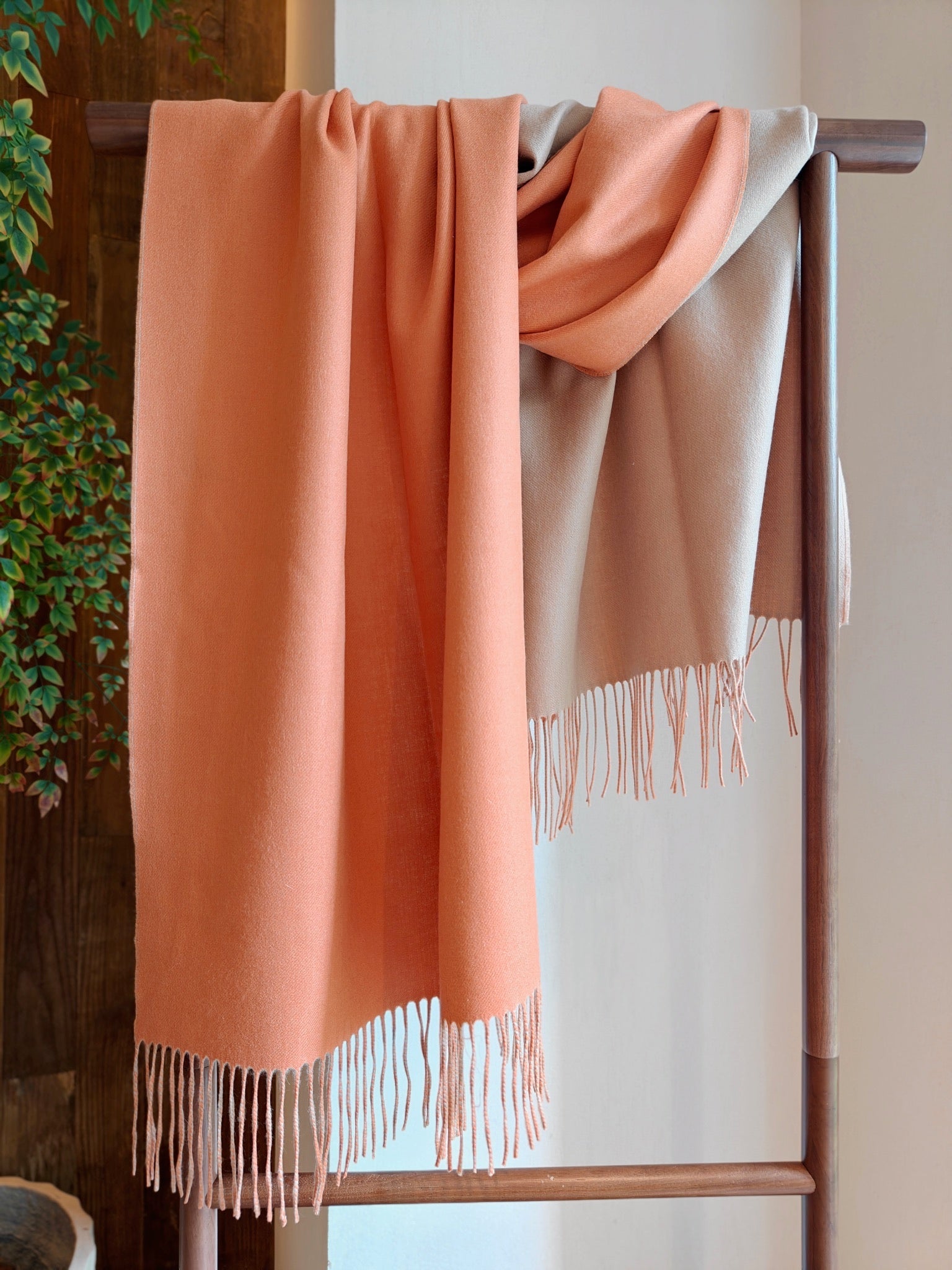 A full draped view of the coral and beige reversible cashmere wrap displayed on a wooden rack. The image highlights the wrap’s silky texture, fluid movement, and soft fringe detailing.
