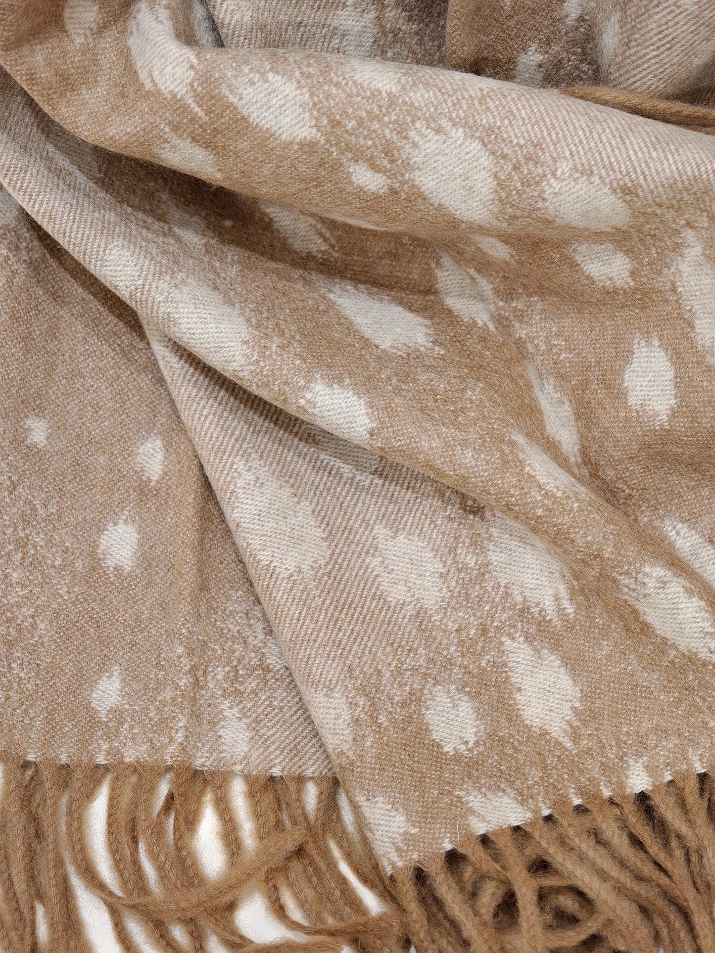 A detailed close up of the fawn cashmere throw showing its soft brushed texture, subtle deer dotted pattern, and long twisted fringe. The photo highlights the plush feel and artisanal weaving.