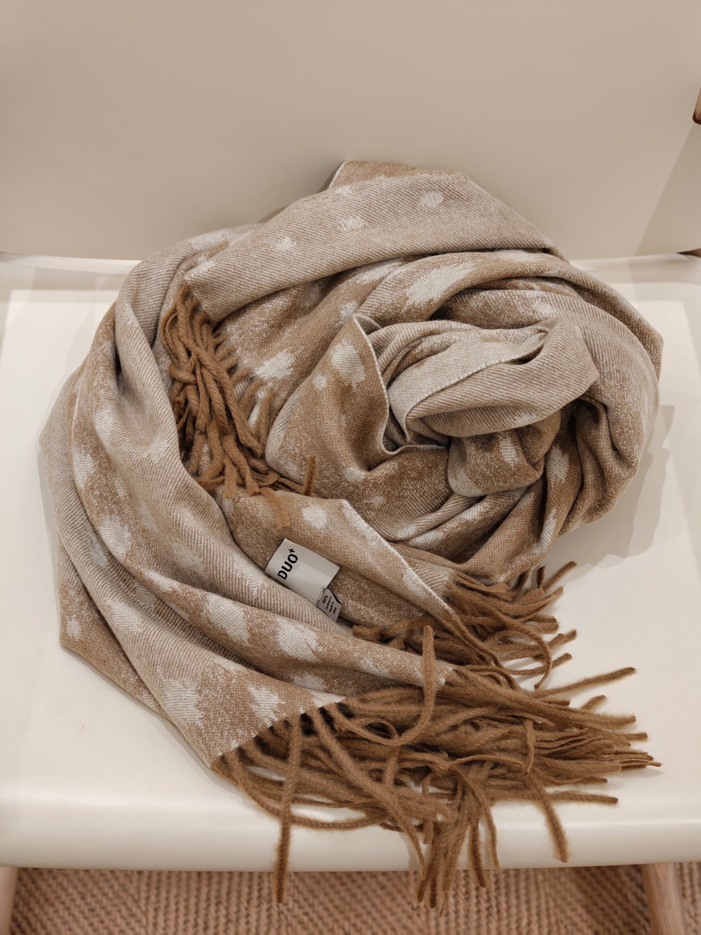 A folded view of the fawn cashmere throw resting on a chair, displaying its airy softness, delicate deer inspired pattern, and plush fringe ends. The image emphasizes comfort and craftsmanship.