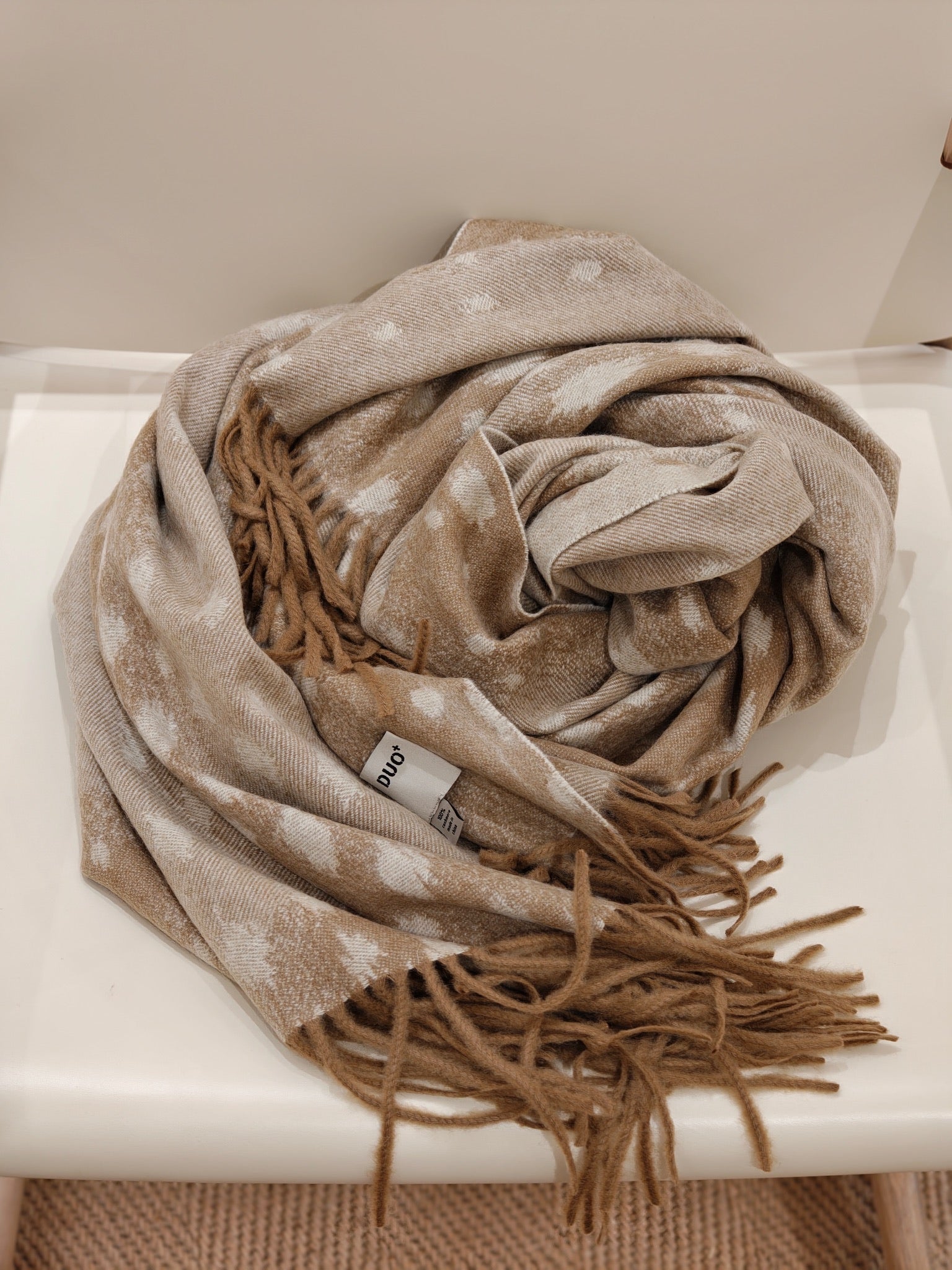 A folded view of the fawn cashmere throw resting on a chair, displaying its airy softness, delicate deer inspired pattern, and plush fringe ends. The image emphasizes comfort and craftsmanship.