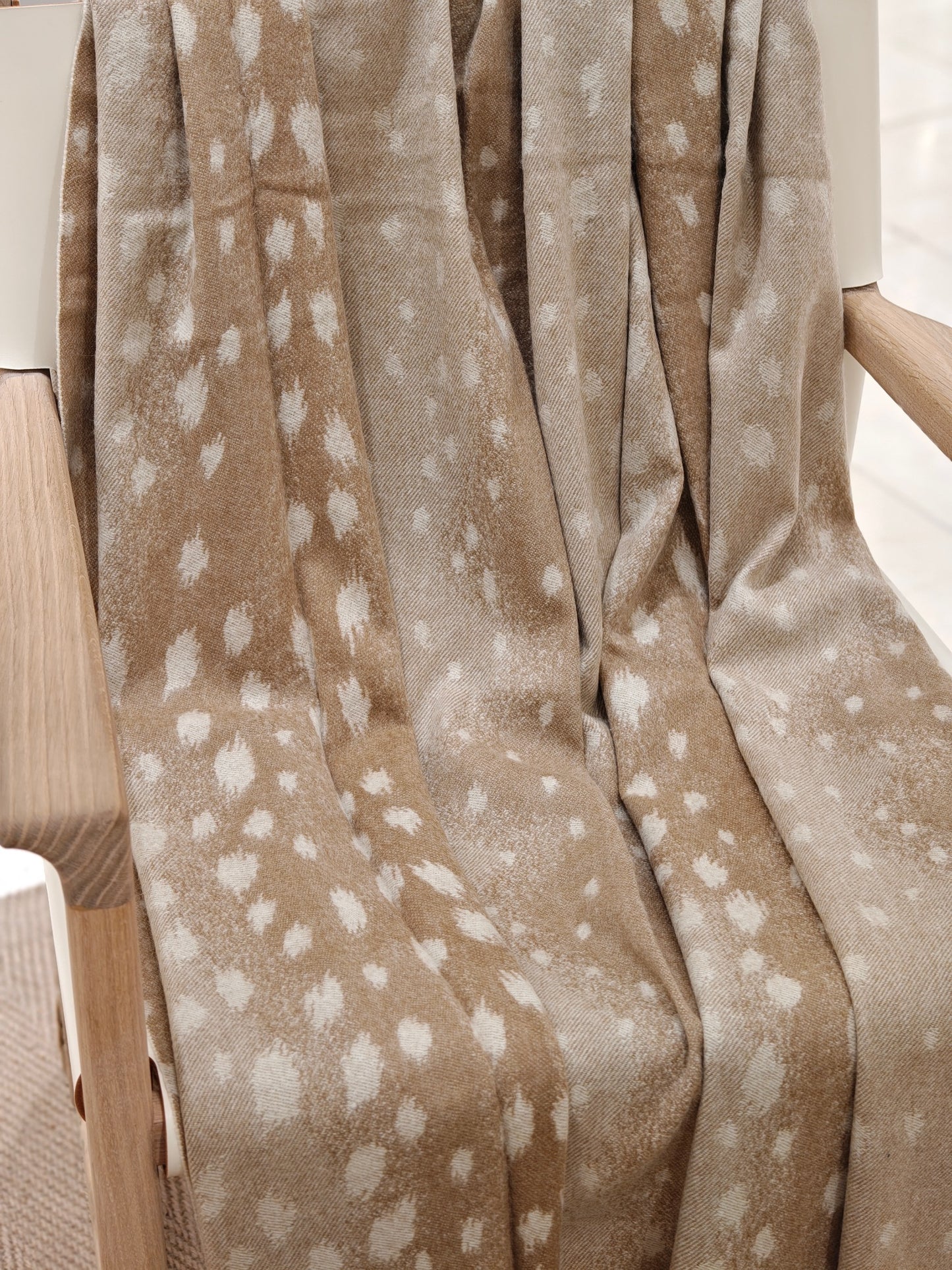 The fawn cashmere throw draped over a wooden chair, revealing its natural dotted pattern and fluid drape. The soft texture and warm neutral tones create a cozy, elevated home aesthetic.