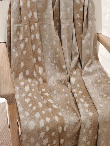 The fawn cashmere throw draped over a wooden chair, revealing its natural dotted pattern and fluid drape. The soft texture and warm neutral tones create a cozy, elevated home aesthetic.