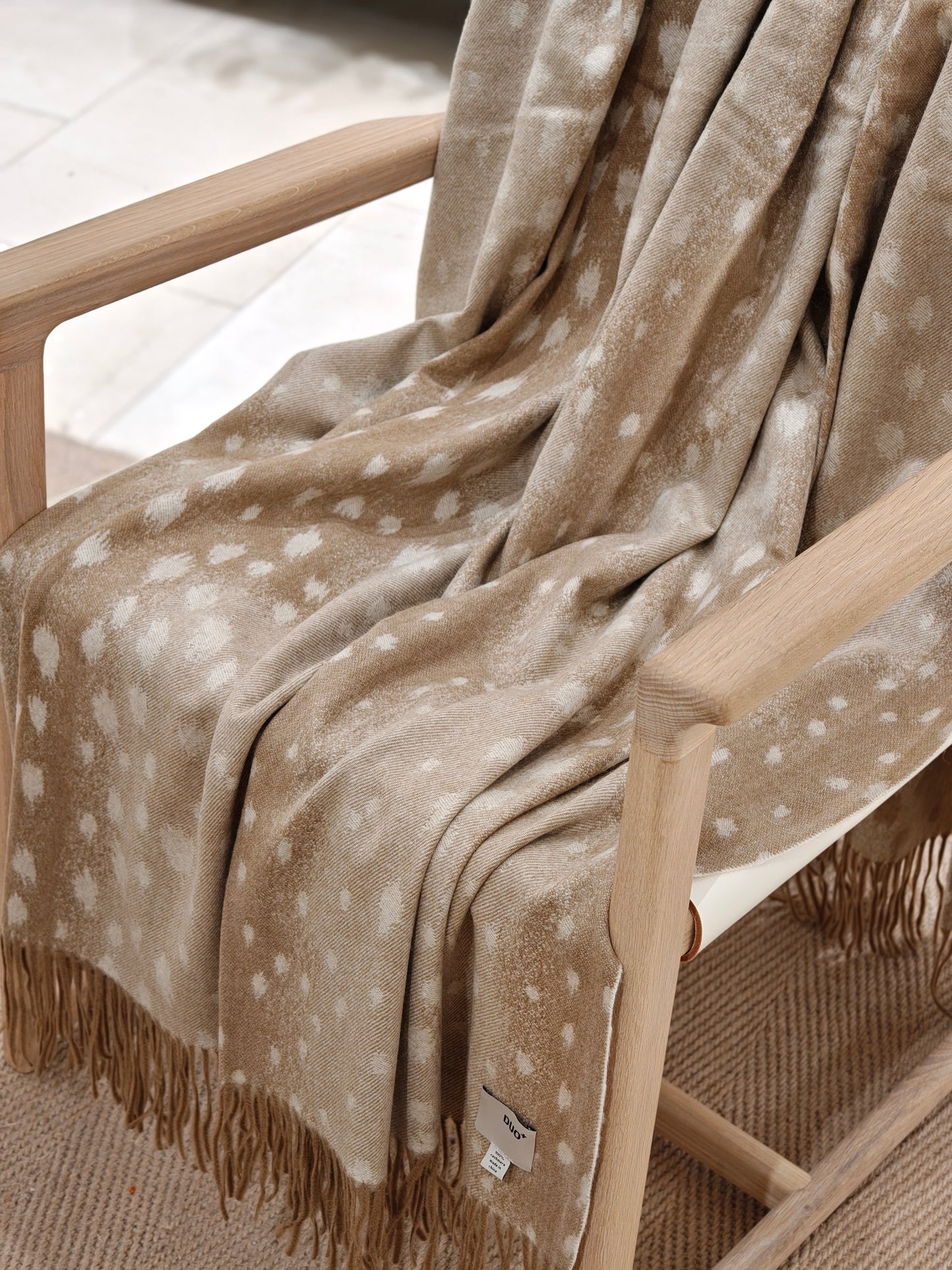 The fawn cashmere throw draped over a wooden chair, revealing its natural dotted pattern and fluid drape. The soft texture and warm neutral tones create a cozy, elevated home aesthetic.