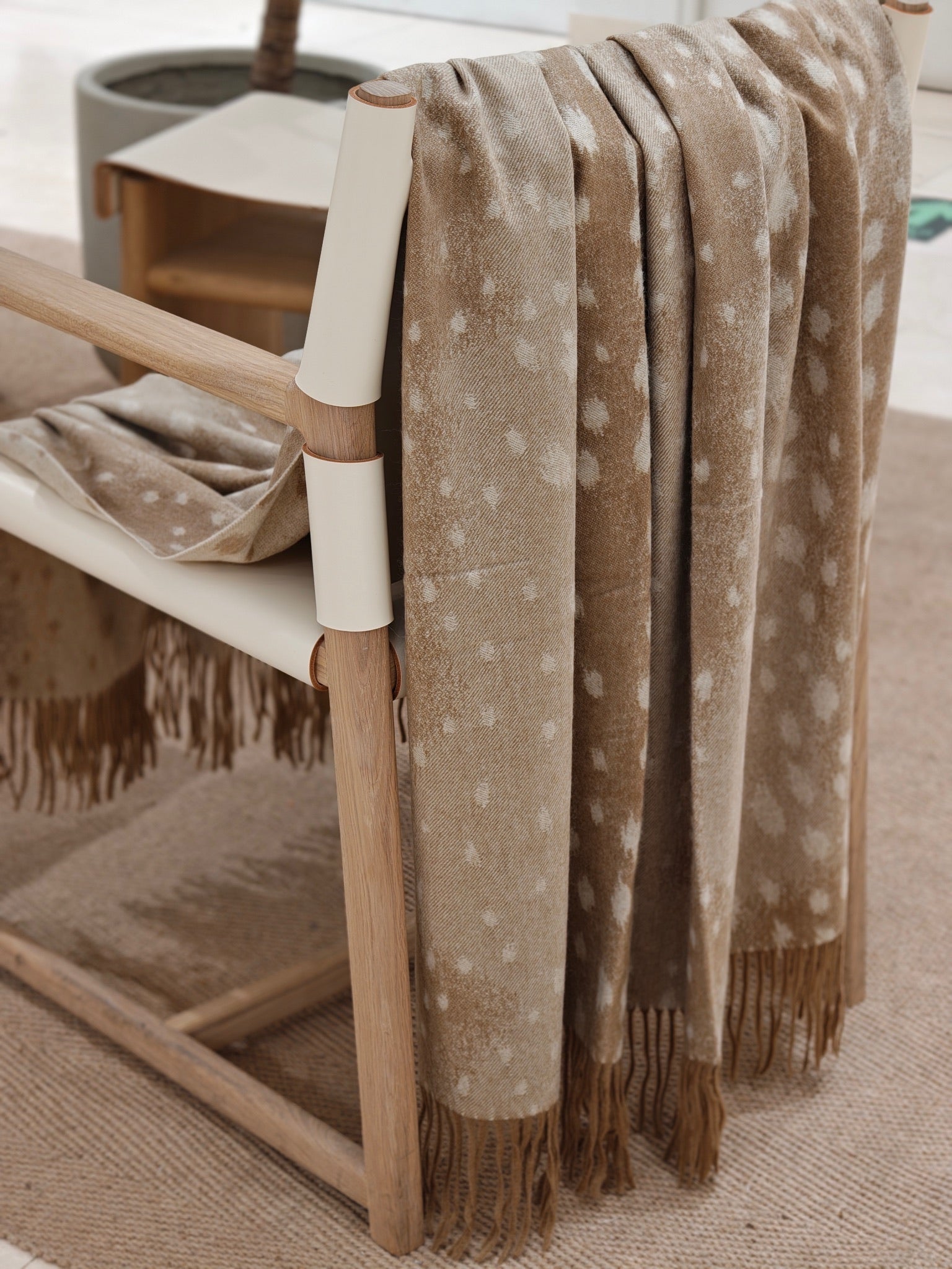 The fawn cashmere throw layered neatly over the back of a wooden chair, showing its soft folds, refined dotted motif, and natural cashmere sheen. The image conveys a cozy, elevated living space.