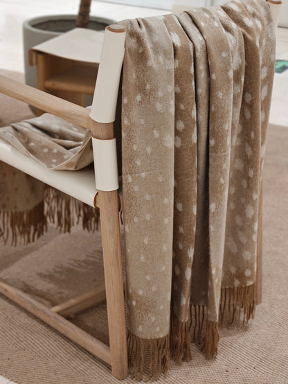 The fawn cashmere throw layered neatly over the back of a wooden chair, showing its soft folds, refined dotted motif, and natural cashmere sheen. The image conveys a cozy, elevated living space.