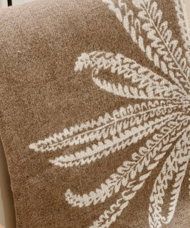A close up of the wool throw showing the intricate white botanical motif against the warm fawn base. The detailed weave emphasizes its softness and artisanal finish.