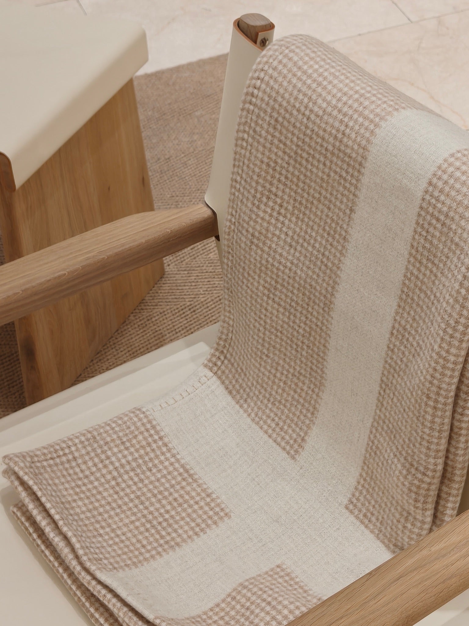 The fawn wool throw draped over a wooden chair, showing its refined houndstooth pattern and smooth drape. The soft beige palette creates a calm and elevated home look.