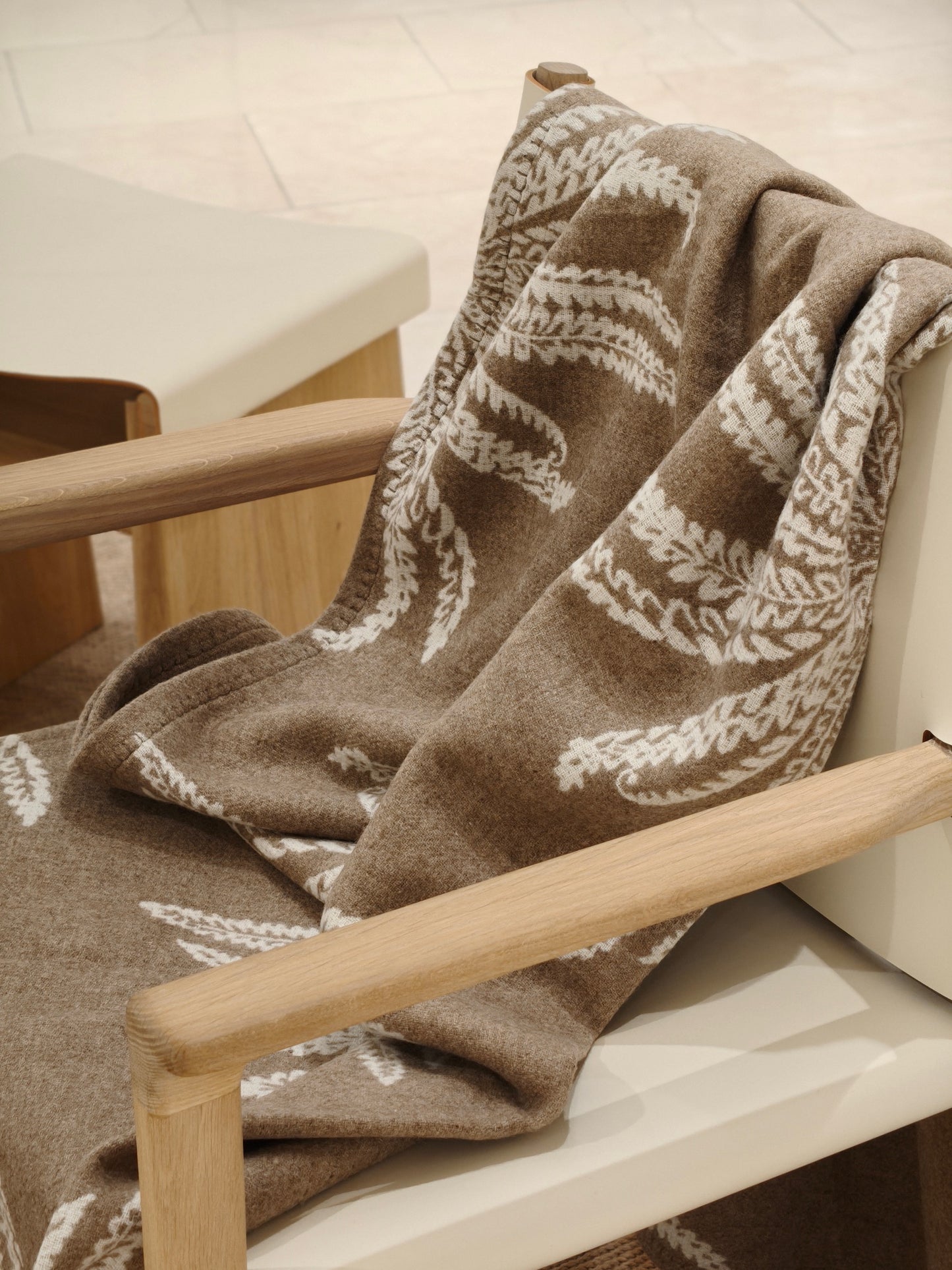 A fawn wool throw draped over a wooden chair, showcasing its soft brushed texture and white botanical pattern. The image highlights the cozy drape and warm neutral tones.