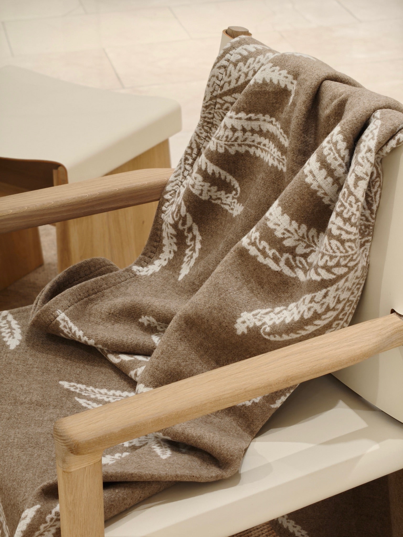 A fawn wool throw draped over a wooden chair, showcasing its soft brushed texture and white botanical pattern. The image highlights the cozy drape and warm neutral tones.