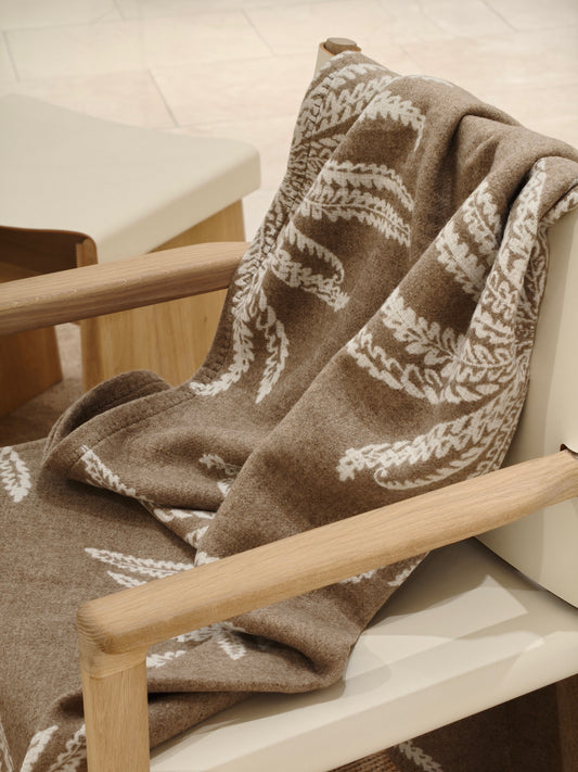 A fawn wool throw draped over a wooden chair, showcasing its soft brushed texture and white botanical pattern. The image highlights the cozy drape and warm neutral tones.