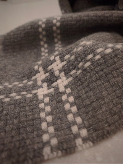 A draped detail shot of the gray cashmere wrap with ivory plaid lines, showing the natural softness and structure of the woven cashmere. The fabric’s surface texture and gentle movement are clearly visible.