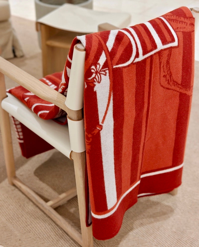 A red equestrian inspired wool throw draped over a modern wooden armchair, showcasing its bold stripes, saddle motifs, and soft woven texture for an elevated home look.
